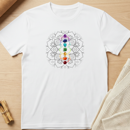 A white short-sleeve crewneck t-shirt laid flat on a light wood surface, featuring a central graphic of the seven chakras in a vertical rainbow sequence over a gray mandala design. Minimalist yoga apparel by pemuyo.