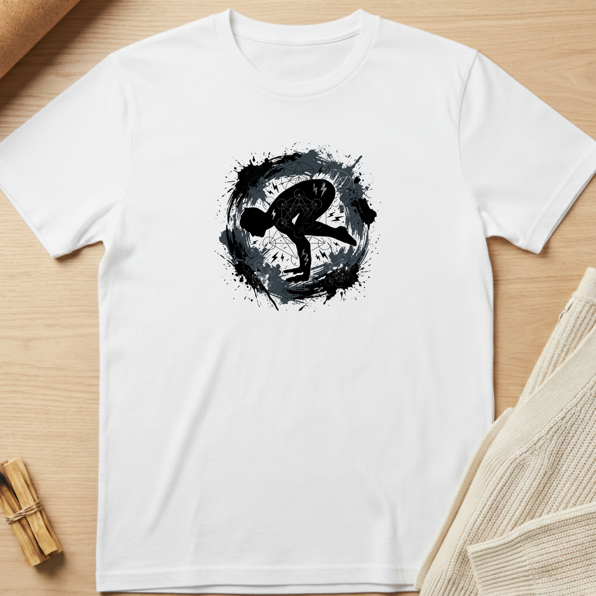 A white short-sleeve crewneck t-shirt laid flat on a light wood surface, featuring a black circular ink-splatter graphic with a stylized human silhouette in the center. Styled flat-lay props include a beige knit fabric at the right and a small wooden bundle at the bottom left. Minimalist yoga apparel by pemuyo.