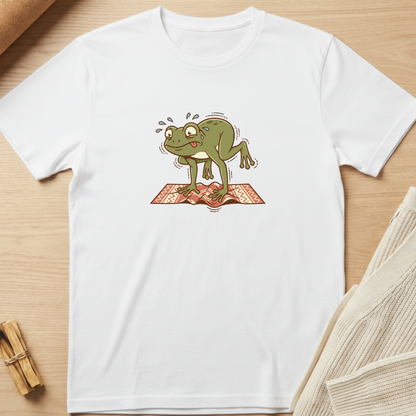 A white short-sleeve crewneck t-shirt laid flat on a light wood surface, featuring a cartoon graphic of a sweating green frog standing on an open book. Minimalist yoga apparel by pemuyo.