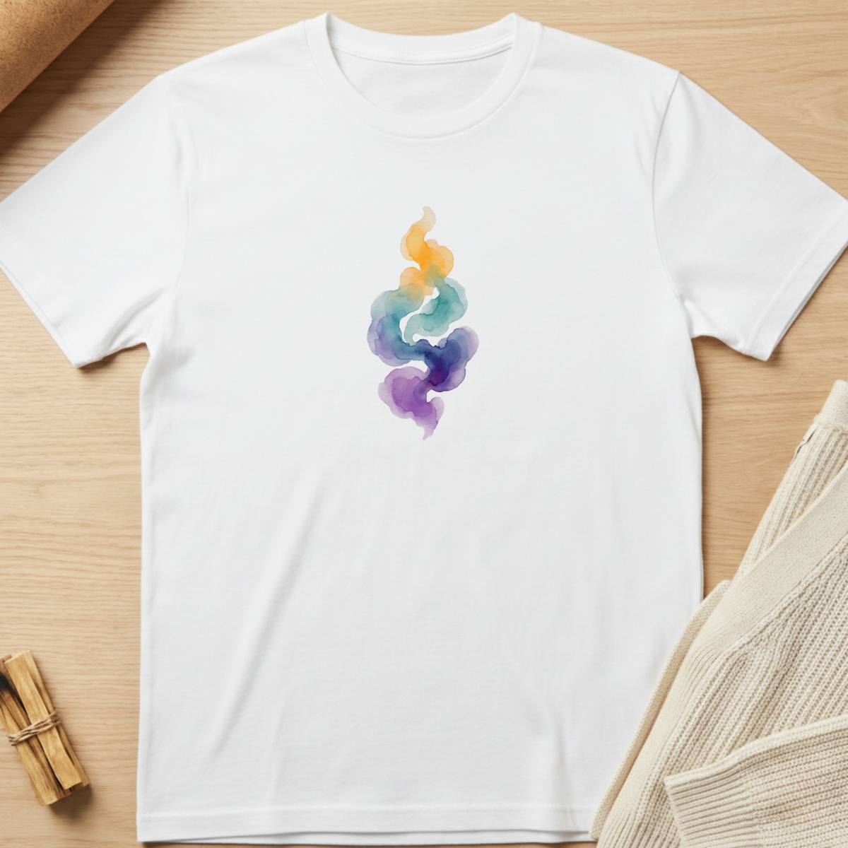 A white short-sleeve crewneck t-shirt with a colorful watercolor smoke graphic in shades of yellow, orange, teal, blue, and purple. The t-shirt is laid flat on a light wood background with styled props, including a beige knit sweater and a small bundle of dried sage leaves. Minimalist yoga apparel by pemuyo.