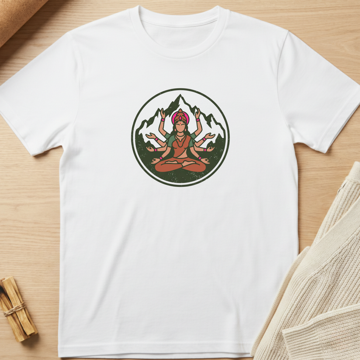 A white short-sleeve crewneck t-shirt laid flat on a light wood surface, featuring a circular graphic of a multi-armed figure meditating in front of mountains. Minimalist yoga apparel by pemuyo.