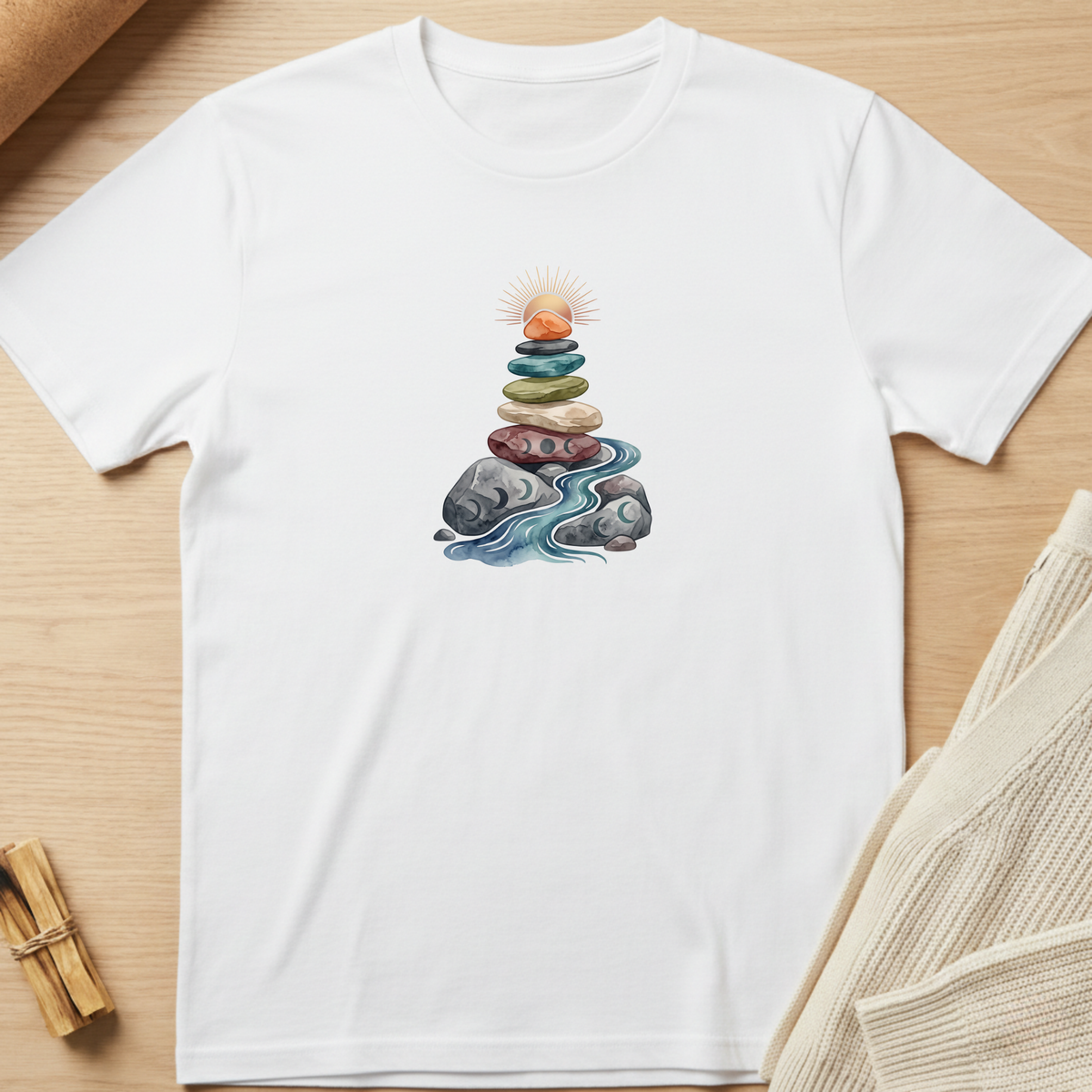 A white short-sleeve crewneck t-shirt laid flat on a light wood background, featuring a central graphic of a stacked stone tower with a sun and blue water. Minimalist yoga apparel by pemuyo.
