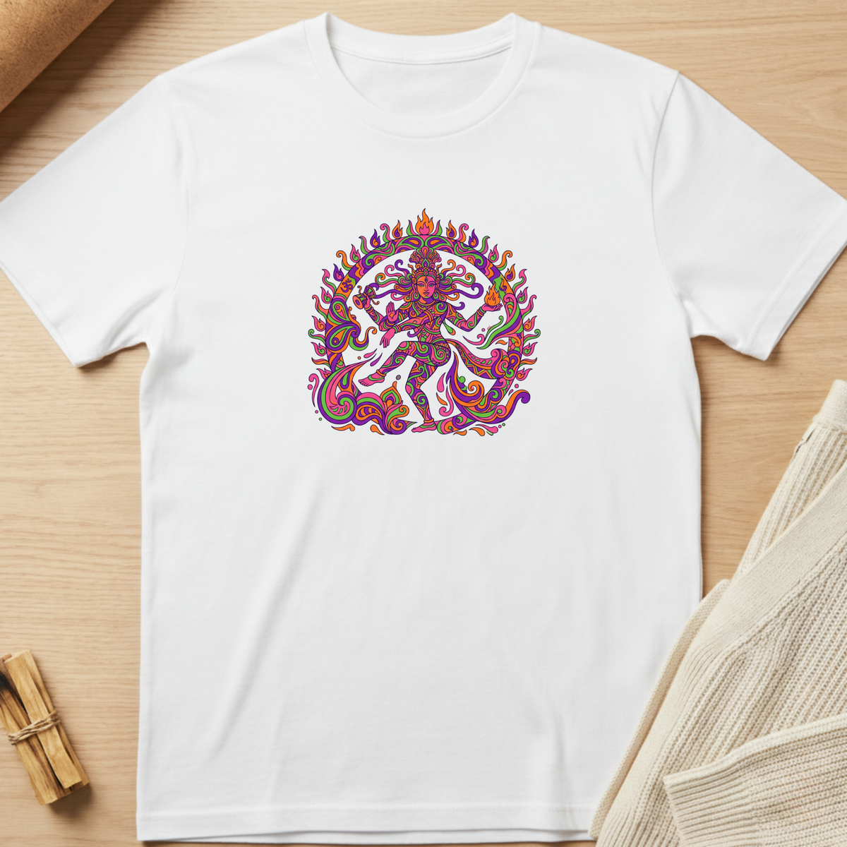 A white short-sleeve crewneck t-shirt laid flat on a light wood surface, featuring a colorful, highly detailed circular graphic of a dancing deity on the chest. Styled flat-lay props include a small bundle of dried herbs in the bottom-left corner and a beige knit fabric in the bottom-right. Minimalist yoga apparel by pemuyo.