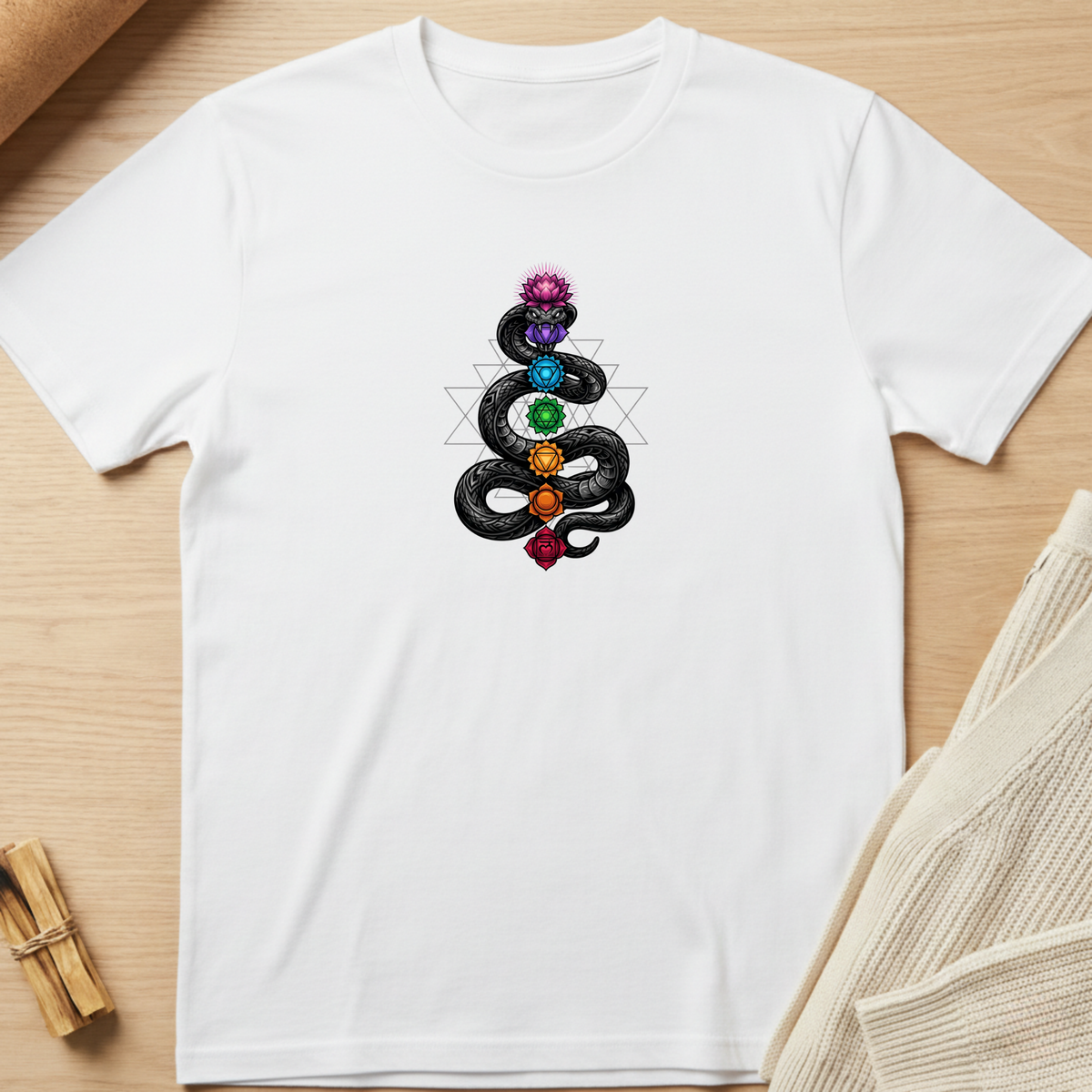A white short-sleeve crewneck t-shirt laid flat on a light wood surface, featuring a central graphic of a black serpent wrapped around a chakra symbol with colorful gemstone-like circles in purple, blue, green, orange, and red. Minimalist yoga apparel by pemuyo.