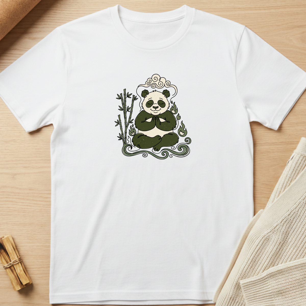 A white short-sleeve crewneck t-shirt laid flat on a light wood surface, featuring a central graphic of a panda sitting cross-legged with bamboo and decorative swirls in dark green and gray. Minimalist yoga apparel by pemuyo.