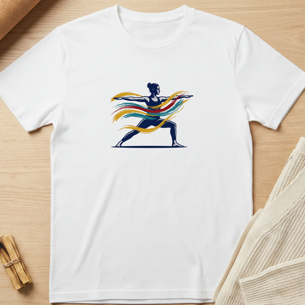 A white short-sleeve crewneck t-shirt laid flat on a light wood surface, featuring a graphic of a yoga warrior silhouette with flowing multicolor ribbons in blue, yellow, orange, and red. Minimalist yoga apparel by pemuyo.