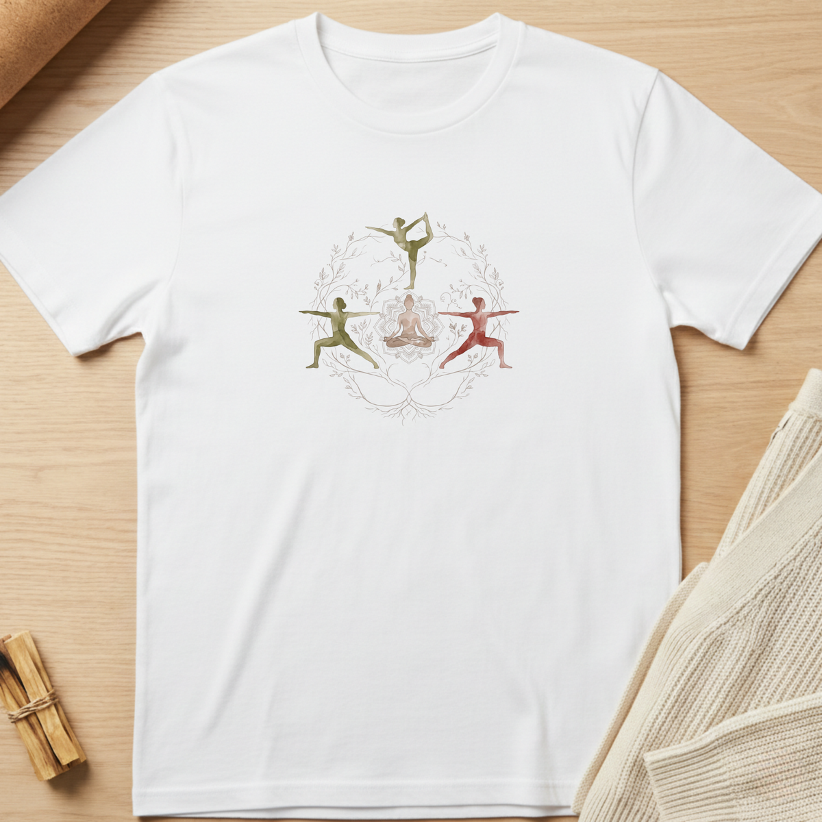 A white short-sleeve crewneck t-shirt laid flat on a light wood surface, featuring a central graphic of three stylized dancers in green, red, and gold within a circular frame. Styled flat-lay props include a beige knit sweater on the right and a small bundle of wooden sticks on the bottom left. Minimalist yoga apparel by pemuyo.