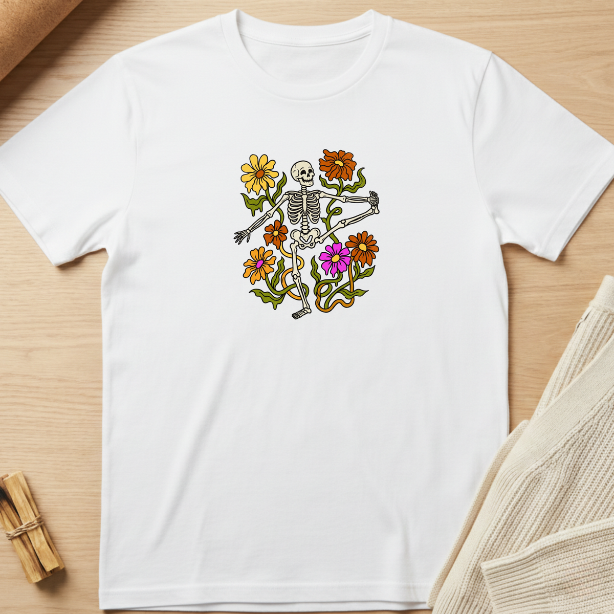 A white short-sleeve crewneck t-shirt laid flat on a light wood surface, featuring a colorful graphic of a skeleton surrounded by yellow, orange, and purple flowers. Minimalist yoga apparel by pemuyo.