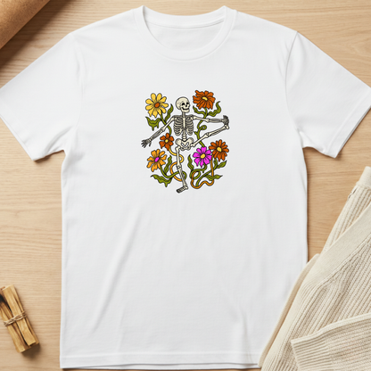 A white short-sleeve crewneck t-shirt laid flat on a light wood surface, featuring a colorful graphic of a skeleton surrounded by yellow, orange, and purple flowers. Minimalist yoga apparel by pemuyo.