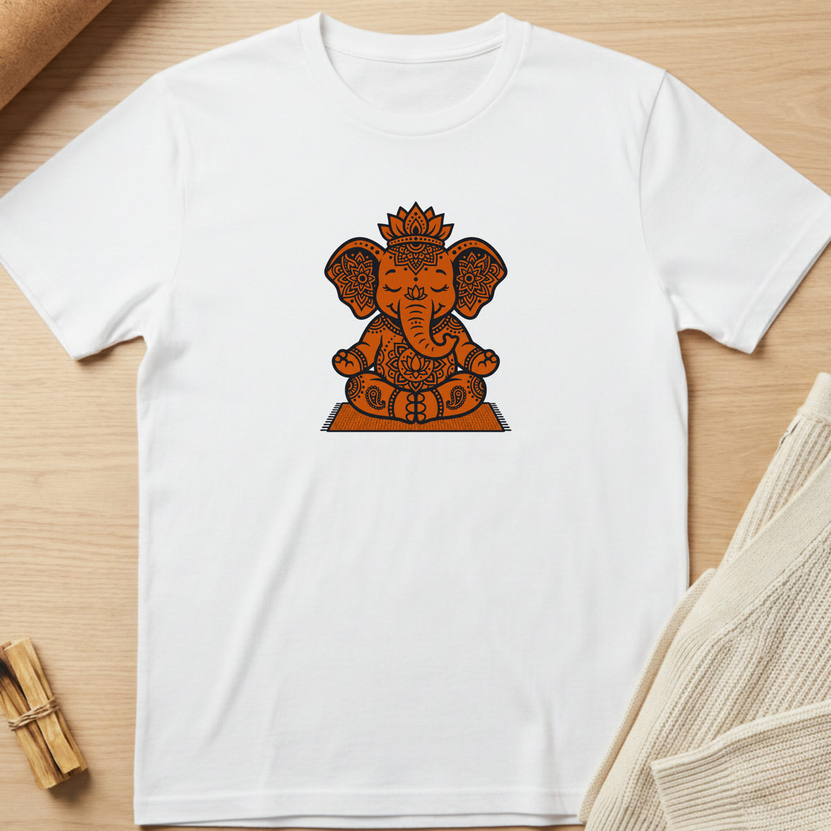 A white short-sleeve crewneck t-shirt featuring a central graphic of Ganesha in orange and brown tones, laid flat on a light wood background with styled props. Minimalist yoga apparel by pemuyo.