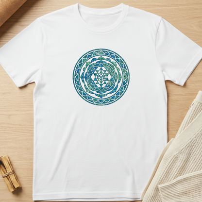 A white short-sleeve crewneck t-shirt featuring a circular mandala-style graphic in shades of blue and green on the chest, laid flat on a light wood background with neutral props. Minimalist yoga apparel by pemuyo.