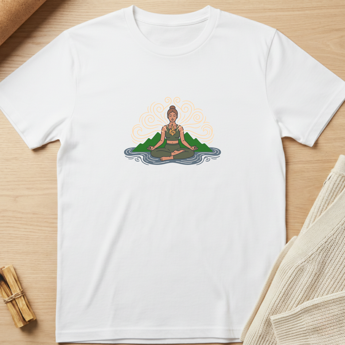 A white short-sleeve crewneck t-shirt laid flat on a light wood surface, featuring a colorful graphic of a woman meditating in front of green mountains and a blue wave, with orange swirls behind her. Minimalist yoga apparel by pemuyo.