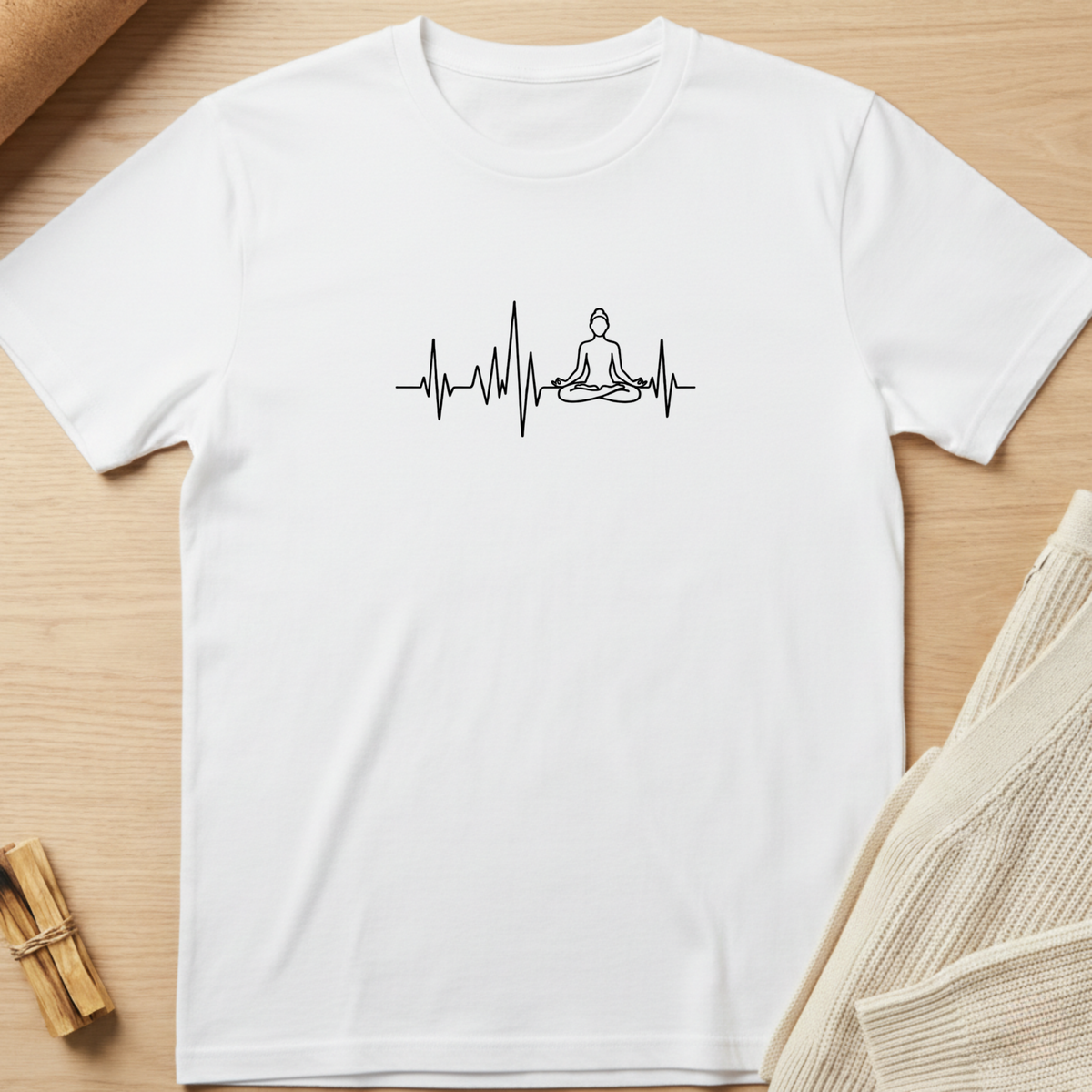 A white short-sleeve crewneck t-shirt laid flat on a light wood background, featuring a black line art graphic of a meditating figure on an EKG-style heartbeat line across the chest. Minimalist yoga apparel by pemuyo.