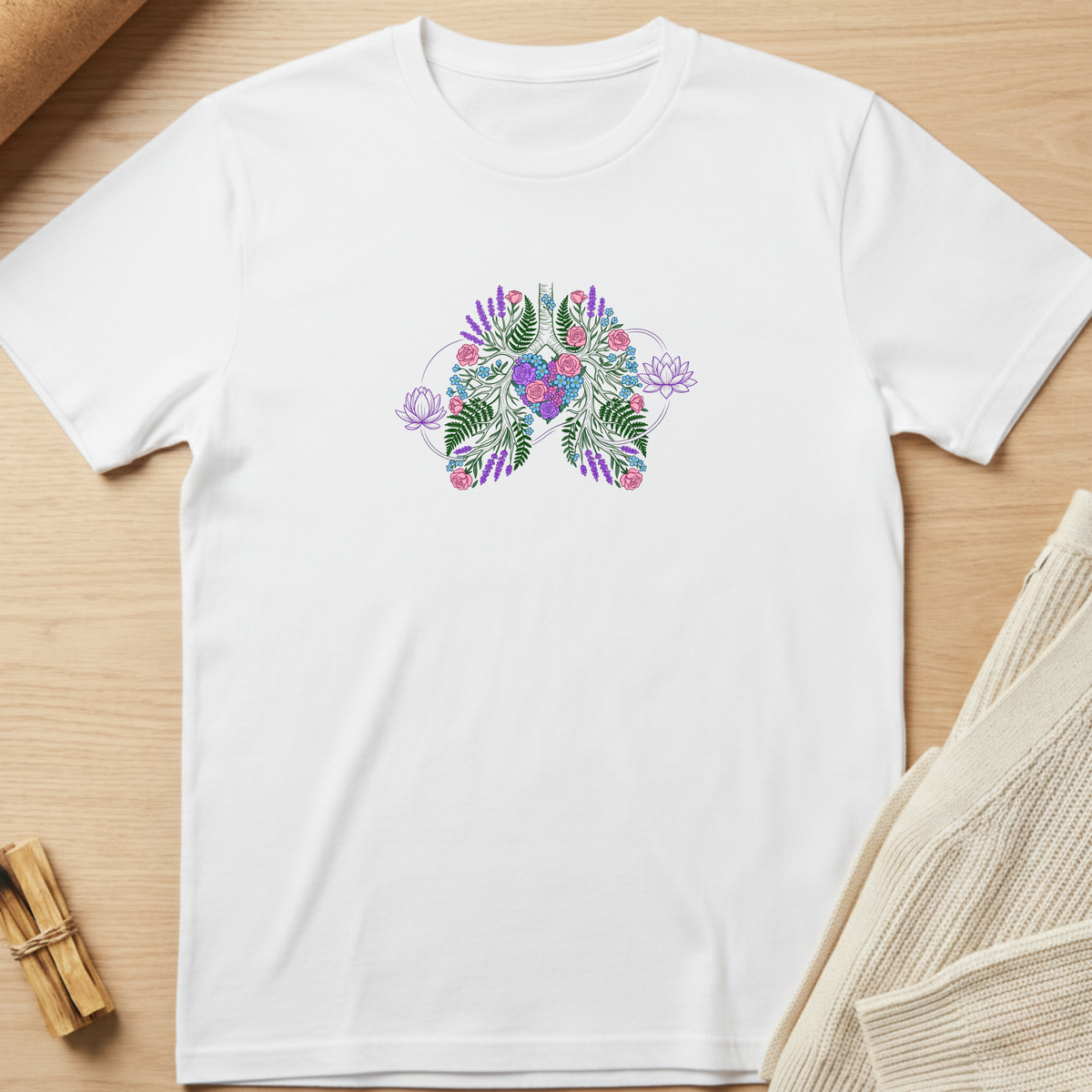 A white short-sleeve crewneck t-shirt laid flat on a light wood surface, featuring a colorful butterfly graphic on the chest made of tropical leaves and flowers in purple, pink, and green. Styled flat-lay props include a rolled brown paper corner, a small bundle of wooden sticks, and a beige knit fabric in the corners. Minimalist yoga apparel by pemuyo.