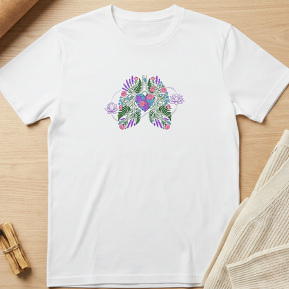 A white short-sleeve crewneck t-shirt laid flat on a light wood surface, featuring a colorful butterfly graphic on the chest made of tropical leaves and flowers in purple, pink, and green. Styled flat-lay props include a rolled brown paper corner, a small bundle of wooden sticks, and a beige knit fabric in the corners. Minimalist yoga apparel by pemuyo.
