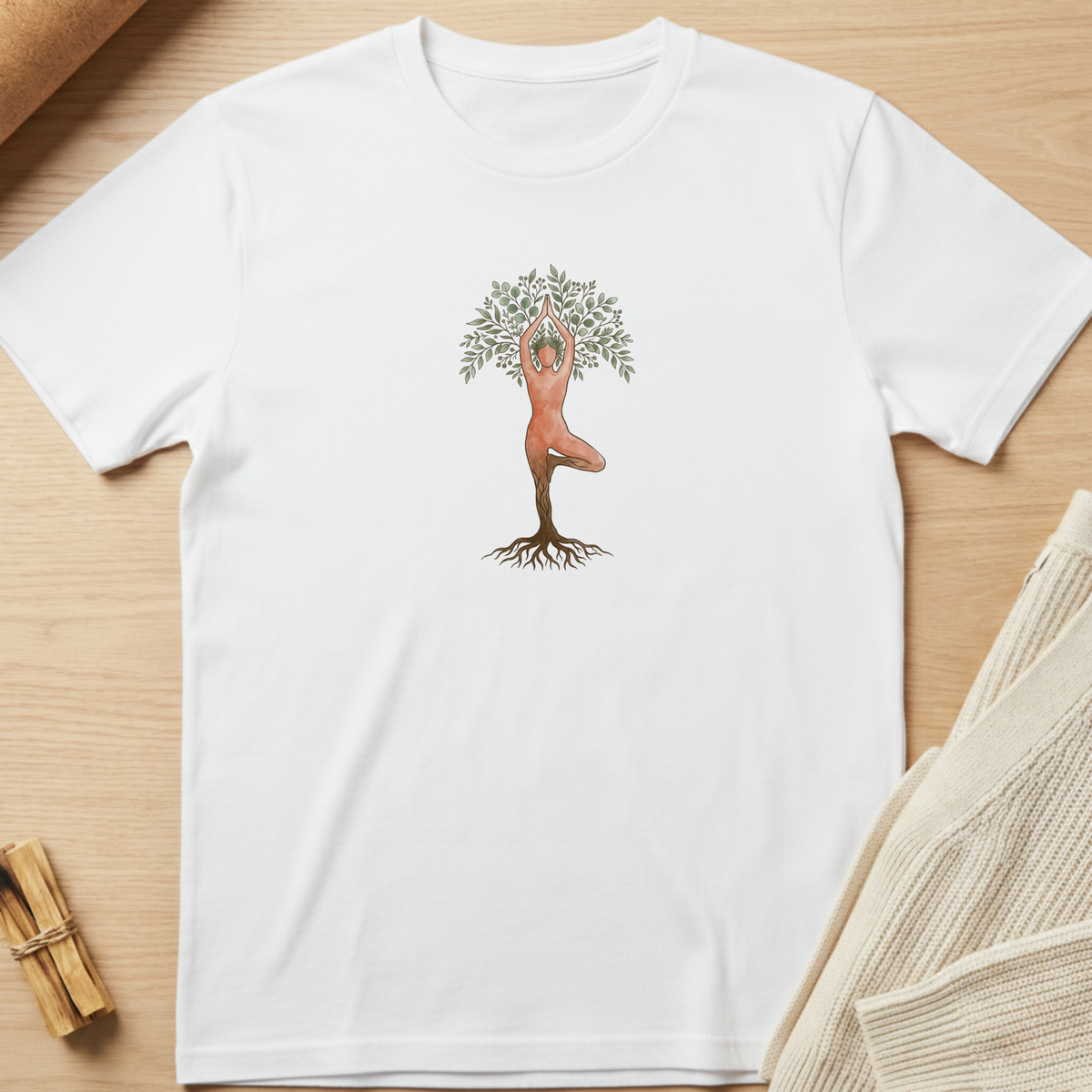 A white short-sleeve crewneck t-shirt laid flat on a light wood surface, featuring a central graphic of a person in a yoga tree pose with green leaves and brown roots. Styled flat-lay props include a beige knit blanket on the right and a small bundle of wooden sticks on the bottom left. Minimalist yoga apparel by pemuyo.