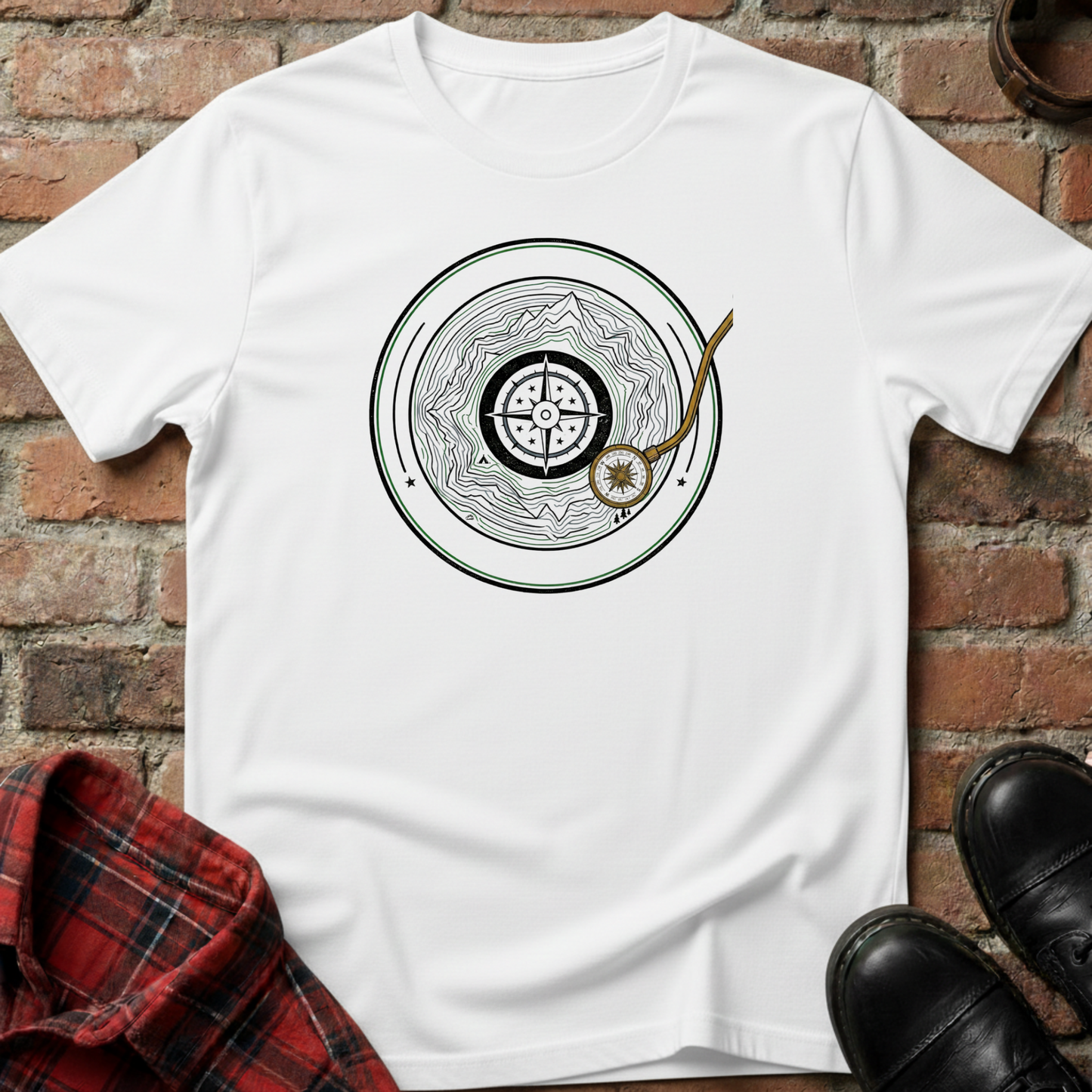 White t-shirt with a circular design featuring a compass and mountain, placed on a brick surface with a plaid shirt and black shoes. Minimalist music apparel by pemuyo.