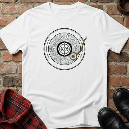 White t-shirt with a circular design featuring a compass and mountain, placed on a brick surface with a plaid shirt and black shoes. Minimalist music apparel by pemuyo.