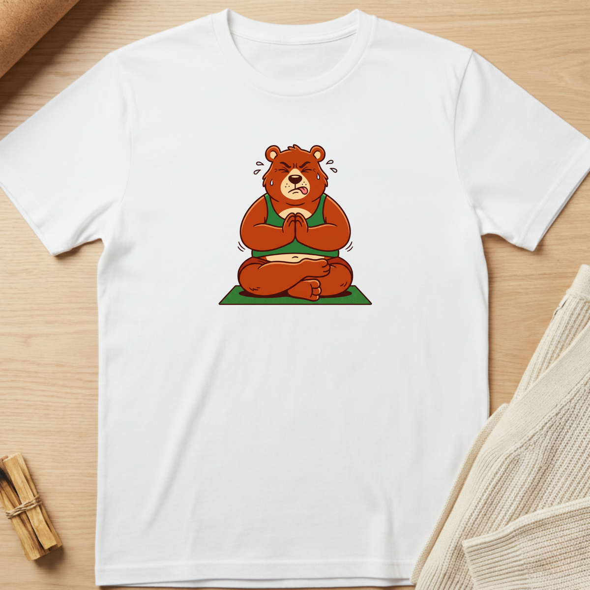 A white short-sleeve crewneck t-shirt laid flat on a light wood surface, featuring a cartoon graphic of a brown bear meditating in a green robe and sitting on a green mat. Minimalist yoga apparel by pemuyo.
