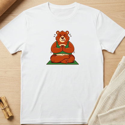 A white short-sleeve crewneck t-shirt laid flat on a light wood surface, featuring a cartoon graphic of a brown bear meditating in a green robe and sitting on a green mat. Minimalist yoga apparel by pemuyo.