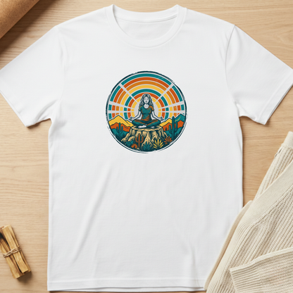 A white short-sleeve crewneck t-shirt laid flat on a light wood surface, featuring a colorful circular graphic of a meditating woman surrounded by mountains and a sunset. Minimalist yoga apparel by pemuyo.